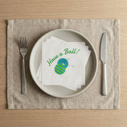 Have a Ball Christmas luncheon napkins stacked on a dinner plate with fork and knife, watercolor holiday party table