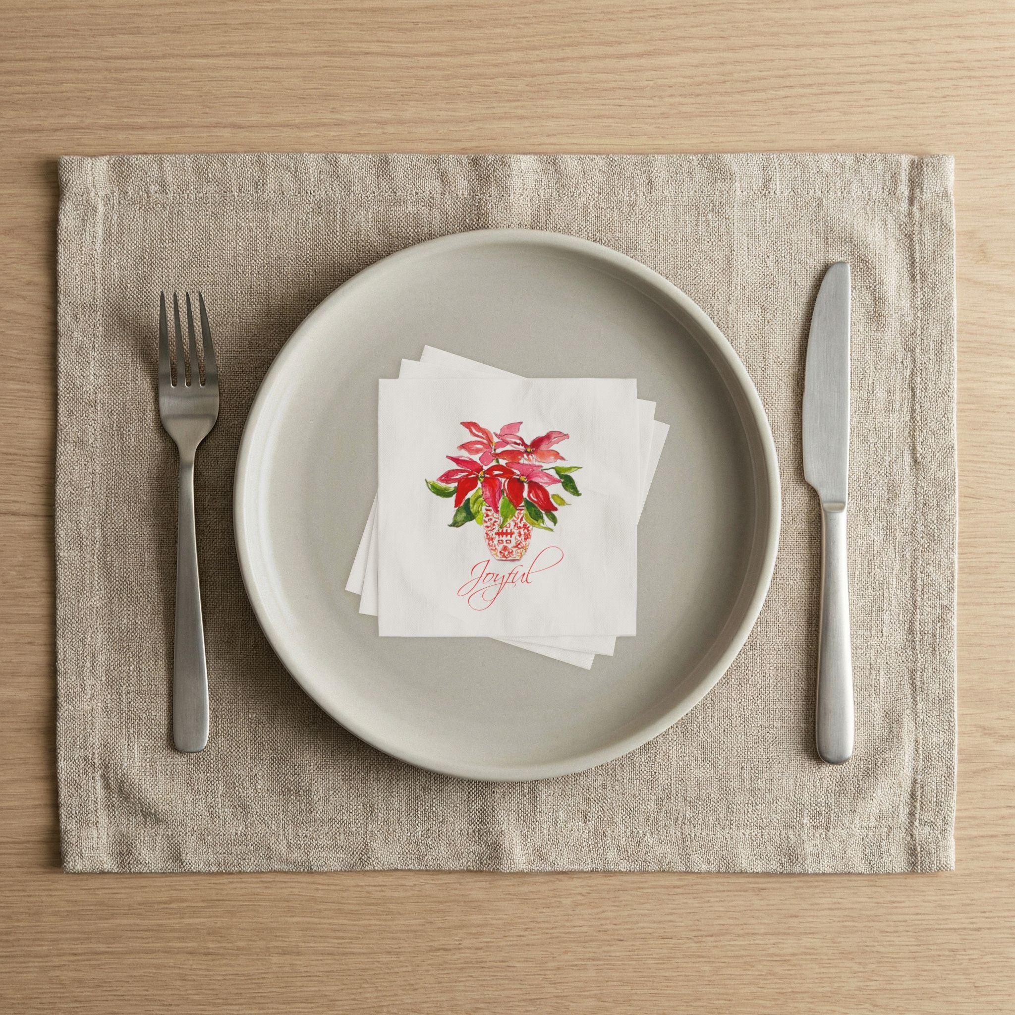 Holiday table place setting with Laura Trevey Joyful poinsettia paper napkins on a linen placemat