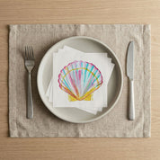 Scallop shell cocktail napkins stacked on a dinner plate with fork and knife, coastal watercolor table setting