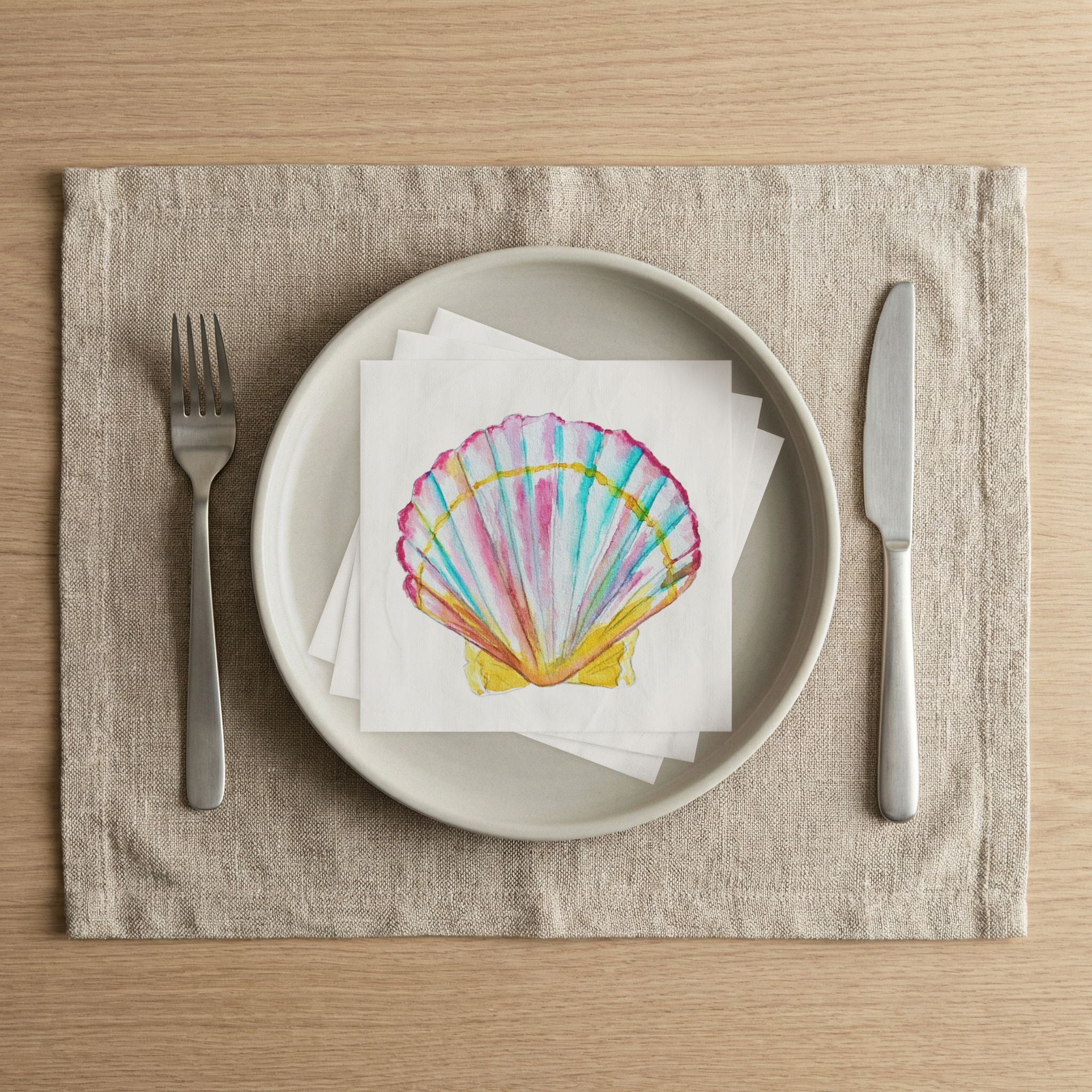 Scallop shell cocktail napkins stacked on a dinner plate with fork and knife, coastal watercolor table setting