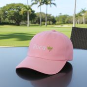 Pink cap with 'Boca' and Palm tree logo on a table with a golf course background