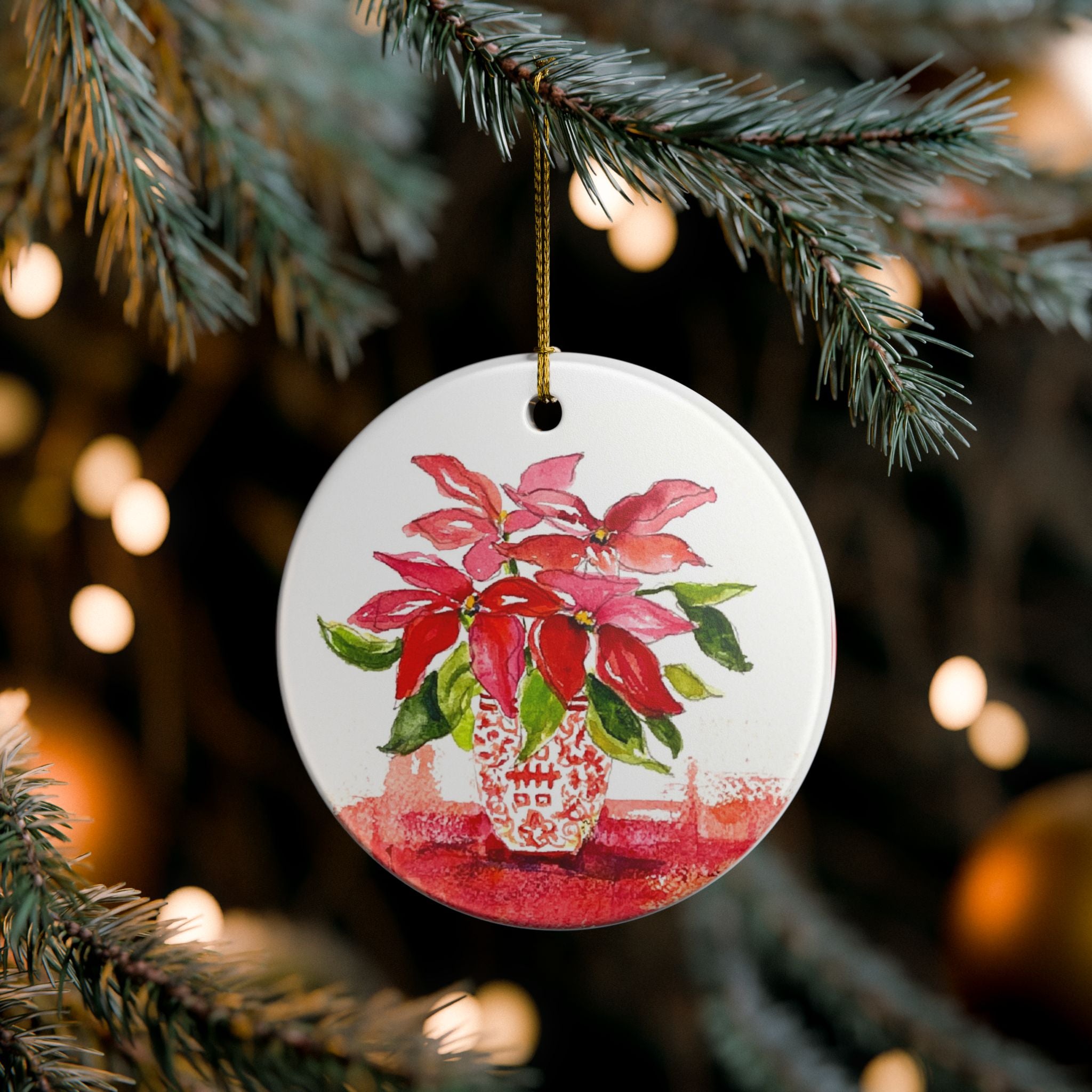 A round white ceramic Christmas ornament featuring a painted red poinsettia in a patterned vase, hanging from a gold string on a pine tree branch with warm bokeh lights in the background.