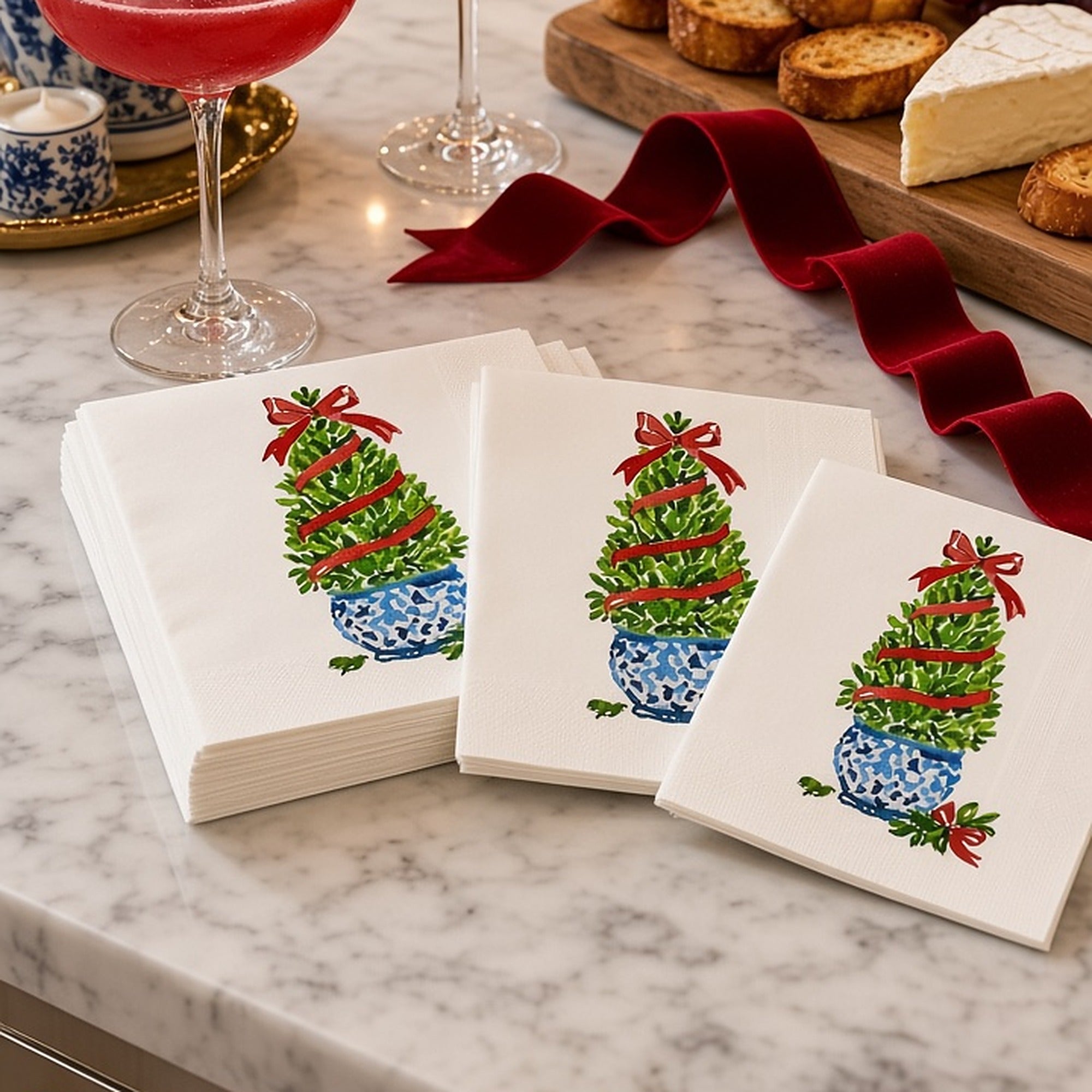 Boxwood Topiary Christmas Cocktail Napkins | Watercolor Holiday Paper Napkins – Boxwood topiary Christmas cocktail napkins by Laura Trevey stacked on a white marble counter with cranberry cocktails, a cheese board and red velvet ribbon