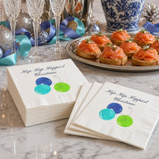 Hap Hap Happiest Christmas Blue Cocktail Napkins | Watercolor Ornament Paper Napkins – Blue Hap Hap Happiest Christmas watercolor napkins by Laura Trevey on white marble with champagne flutes, smoked salmon crostini and white amaryllis in a blue-and-white vase