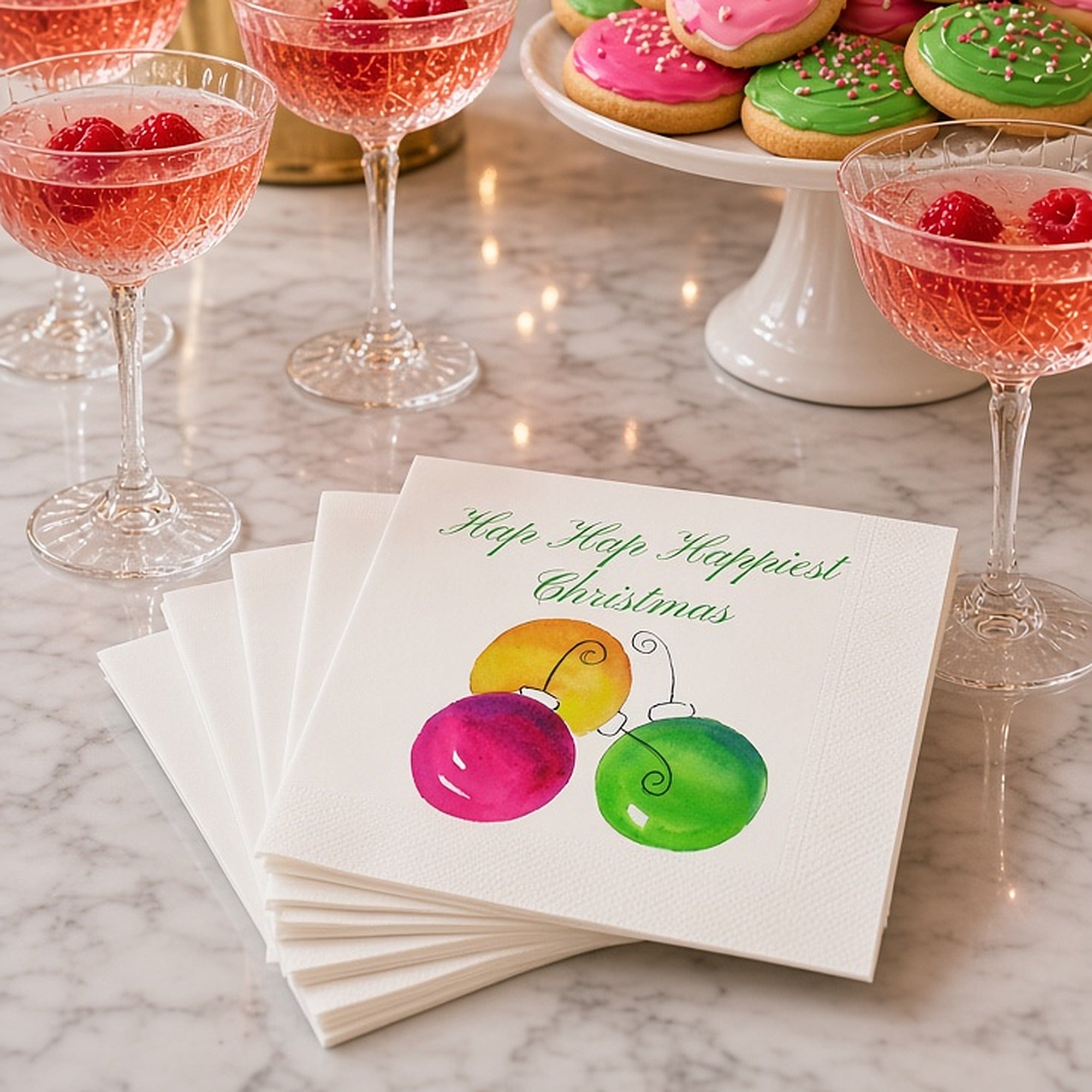 Hap Hap Happiest Christmas Cocktail Napkins | Watercolor Ornament Paper Napkins – Hap Hap Happiest Christmas watercolor ornament napkins by Laura Trevey on white marble with pink champagne coupes, frosted Christmas cookies and a pink and lime tree