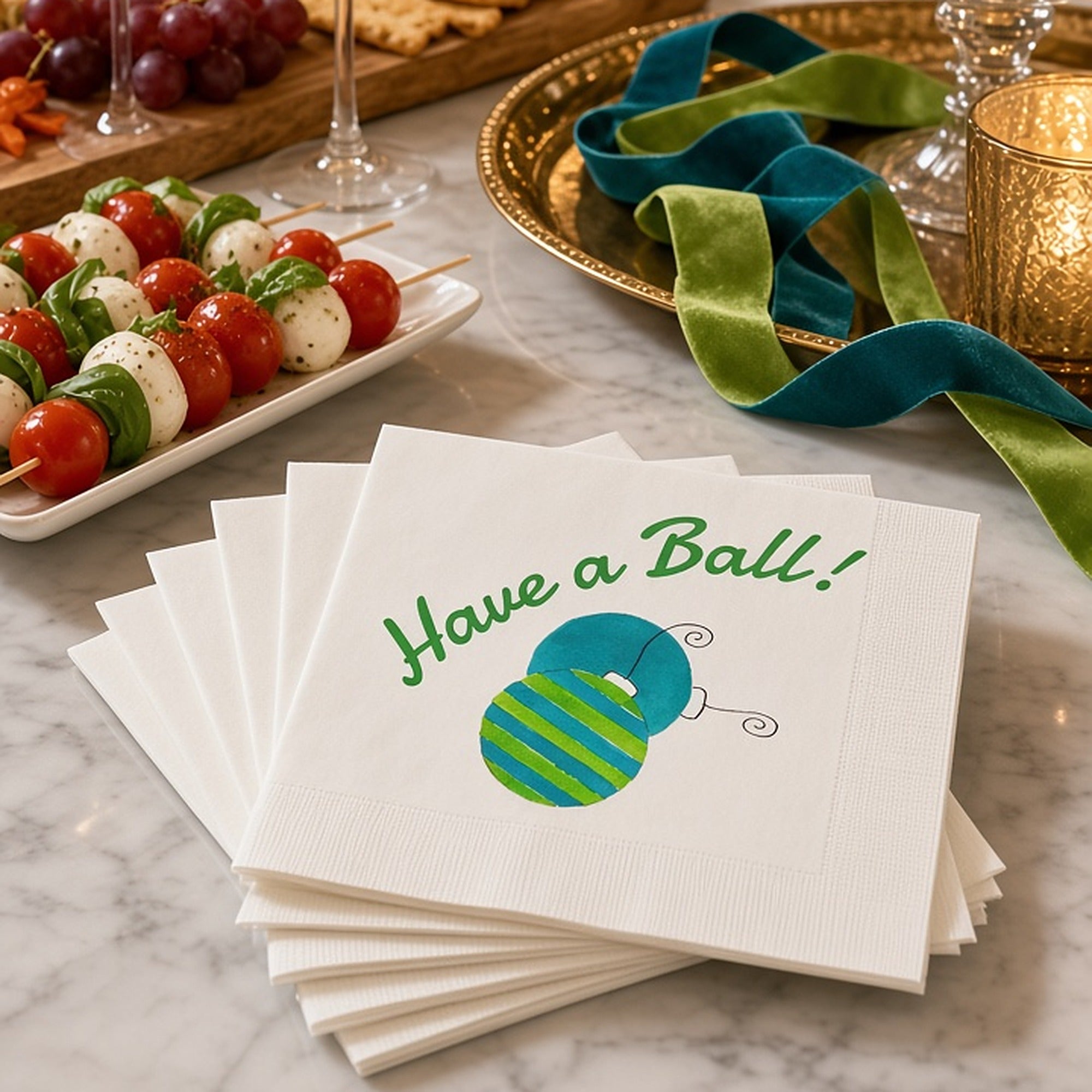 Have a Ball Christmas Cocktail Napkins | Watercolor Ornament Paper Napkins – Stack of Have a Ball watercolor ornament napkins by Laura Trevey on white marble with caprese skewers, a cheese board, sparkling cocktails and teal and lime velvet ribbon