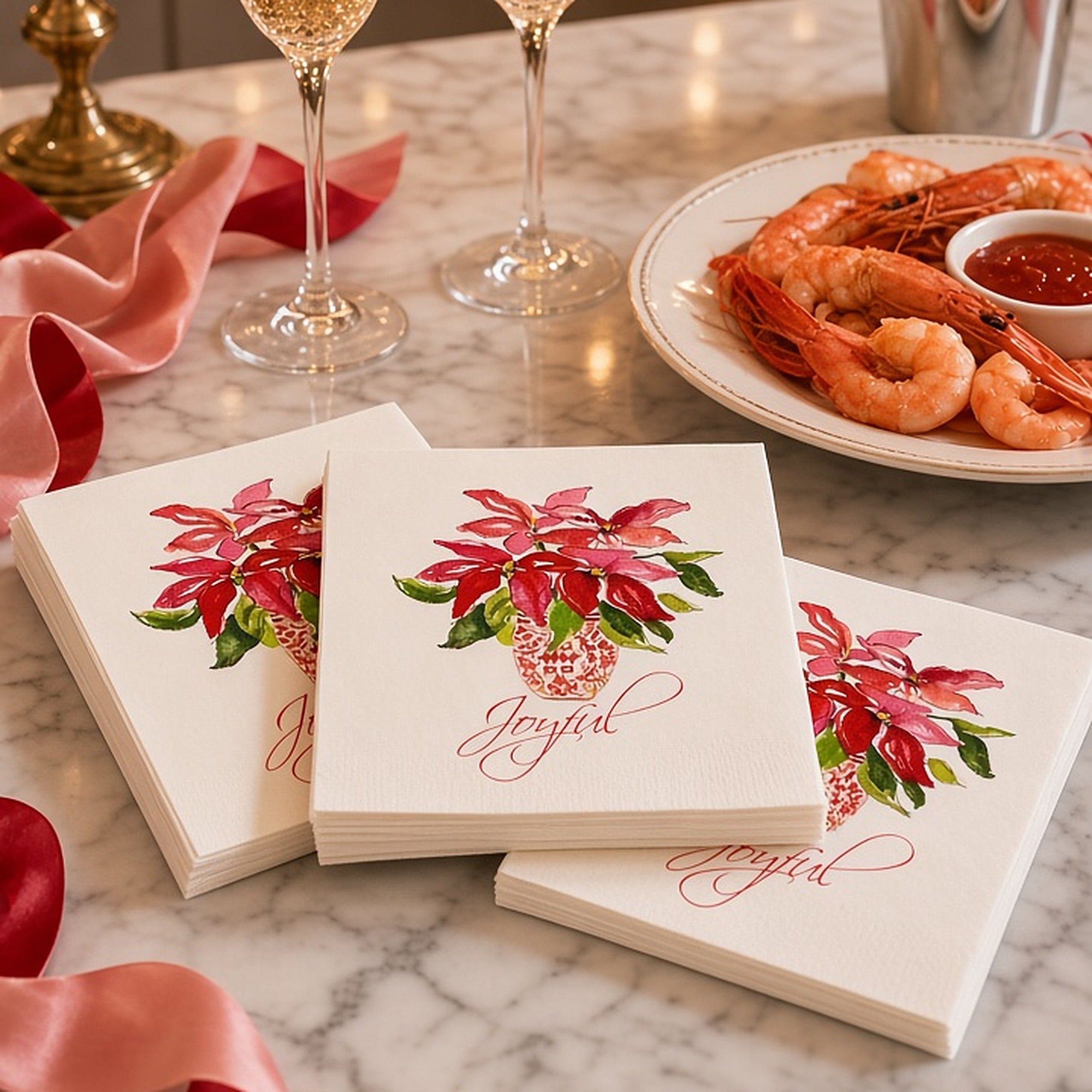 Joyful Poinsettia Christmas Cocktail Napkins | Watercolor Holiday Paper Napkins – Joyful poinsettia Christmas cocktail napkins by Laura Trevey on a white marble counter with champagne flutes, shrimp cocktail and pink and red ribbon